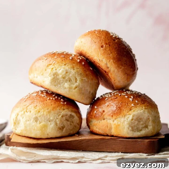 Side view of a stack of fluffy gluten-free hamburger buns with shiny, golden crusts and sesame seed topping, placed on a wooden board with a soft pink background.