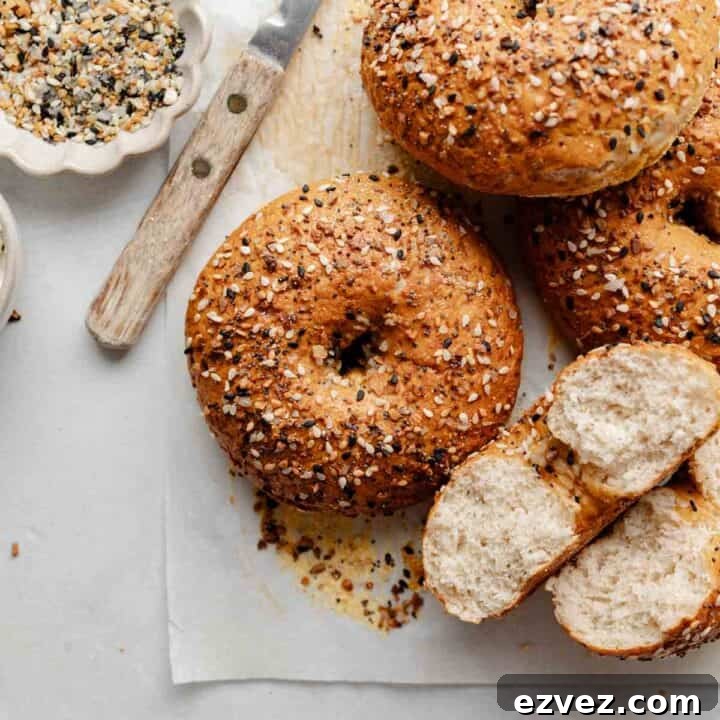 A close-up shot of a gluten-free bagel with a crispy, golden crust covered in everything bagel seasoning, with a knife and small bowl of seasoning nearby. One bagel is cut open to reveal the airy interior.
