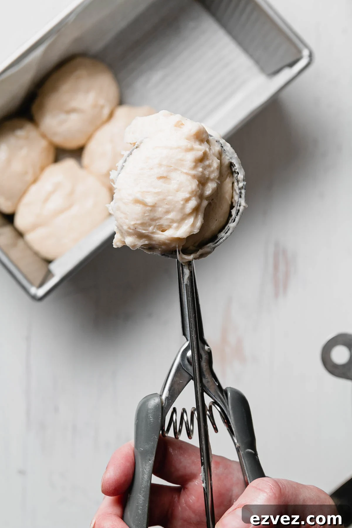A cookie scoop holding sticky gluten-free roll dough above a pan of rolls.