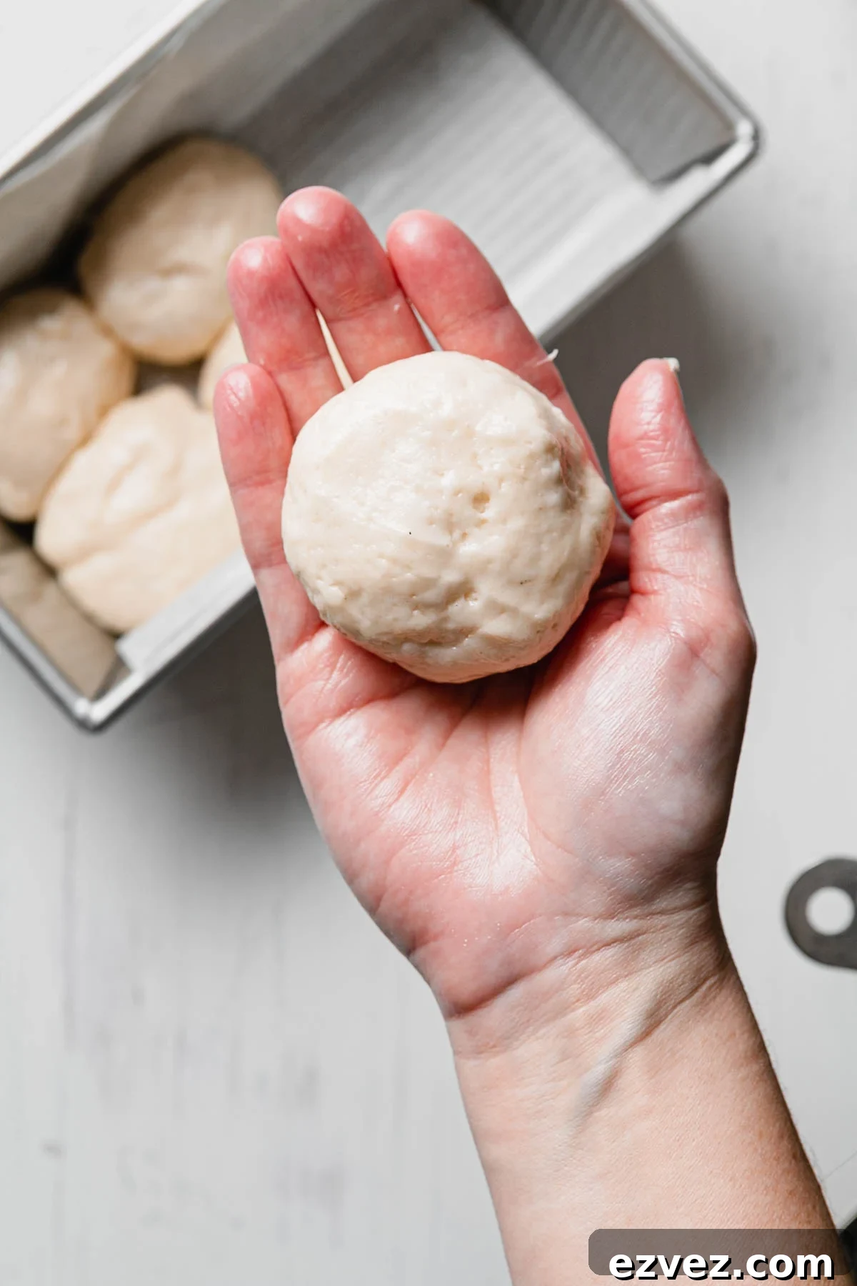 A hand holding a shaped gluten-free Hawaiian roll dough ball over a loaf pan.