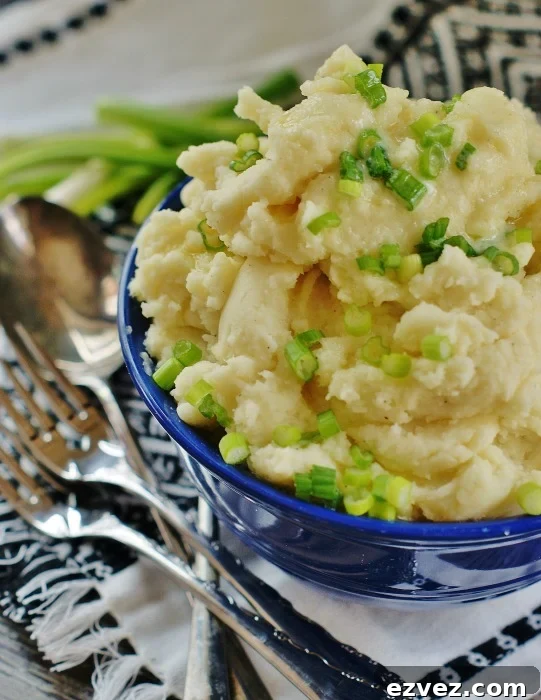 Upclose of a blue Fiestaware serving bowl piled high with horseradish mashed potatoes garnished with chives and drizzled with butter. A white and black tea towel, silverware and green onions are in the background.