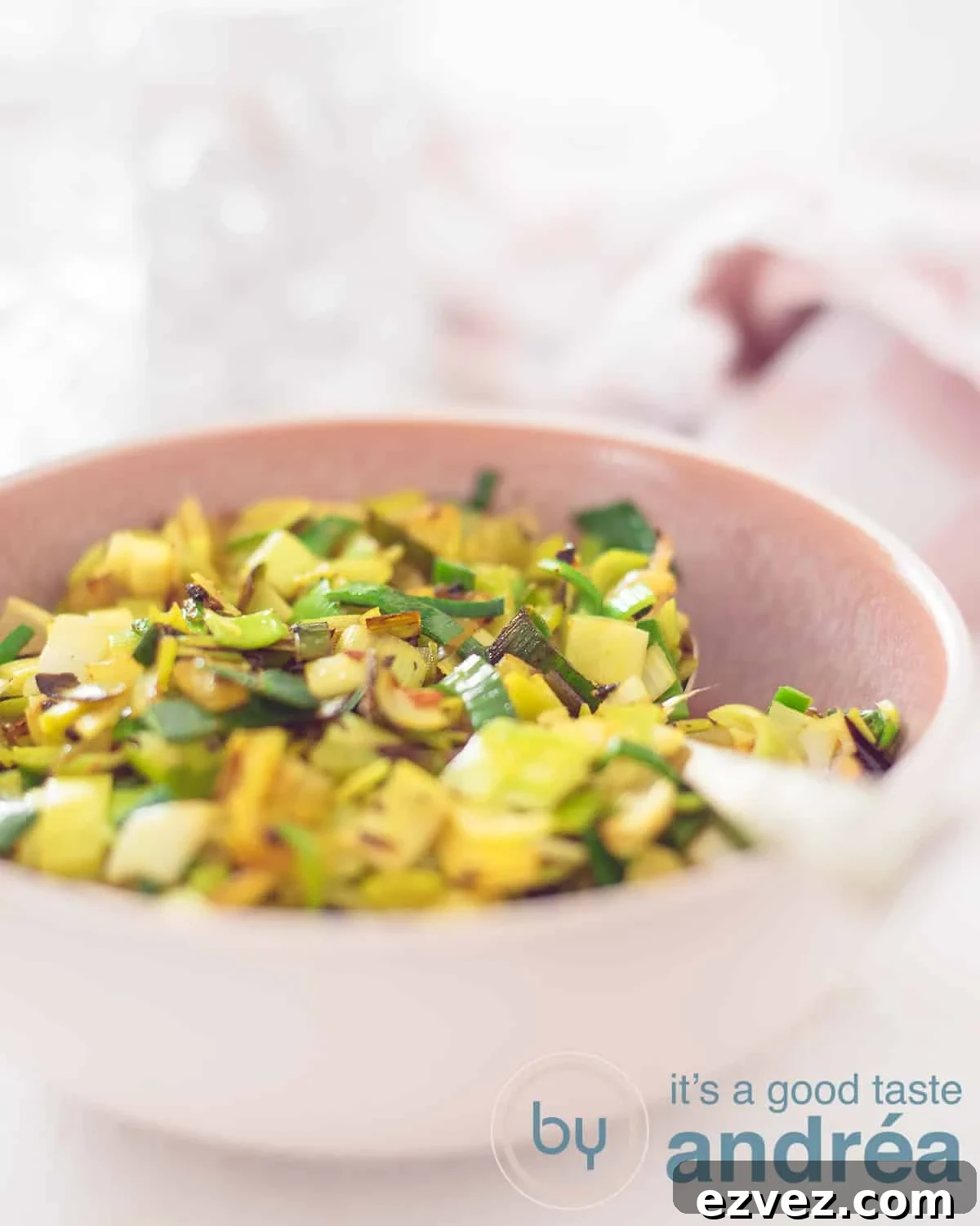 An elevation shot showing part of a pink bowl filled with curry leeks and chilli. A golden spoon sticks into it. A white background, with a glass and a pink and white tea towel in the background.