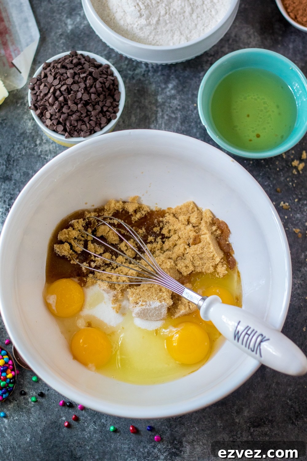 Cosmic Brownies Recipe — Fudgy, No-Bake Chocolate Squares 4 eggs, sugar, and vanilla in a white mixing bowl with a whisk off to the side.