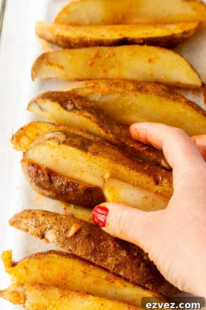 Par-cooked russet potato wedges being stirred with oil and seasoning by hand in a large glass dish.