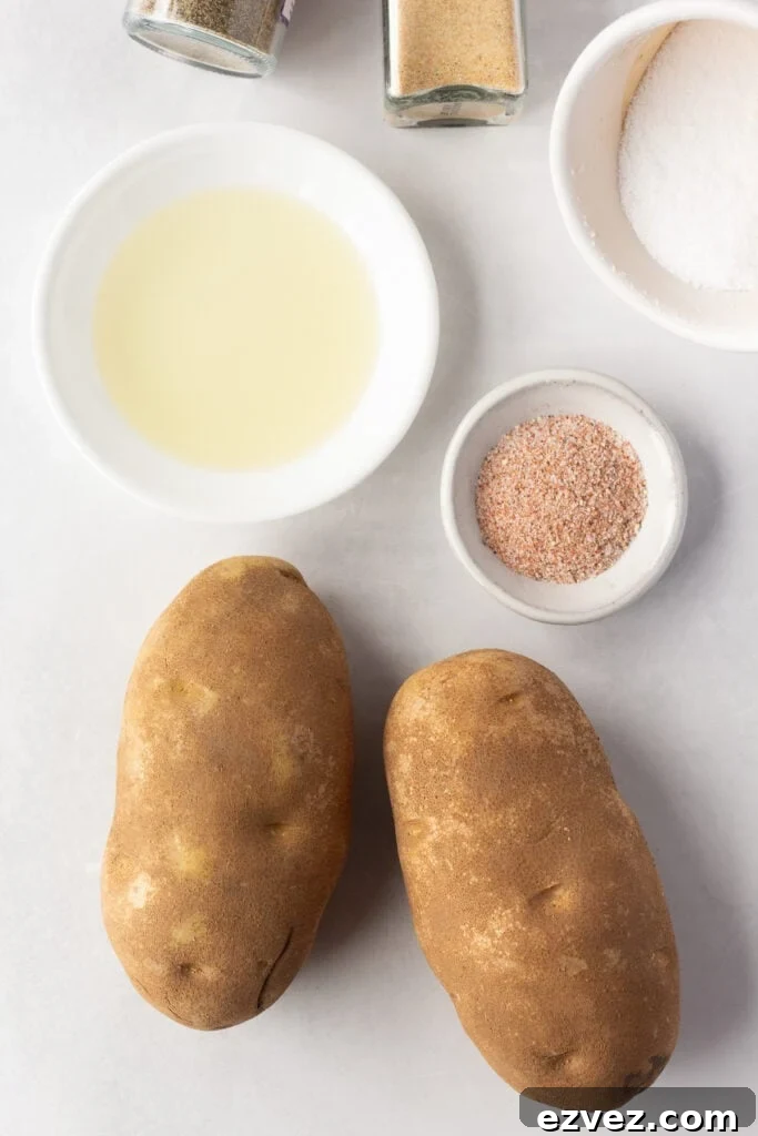 Top down shot of two russet potatoes, a small plate with oil, and a small bowl with a red-ish seasoning on it on a light gray surface.