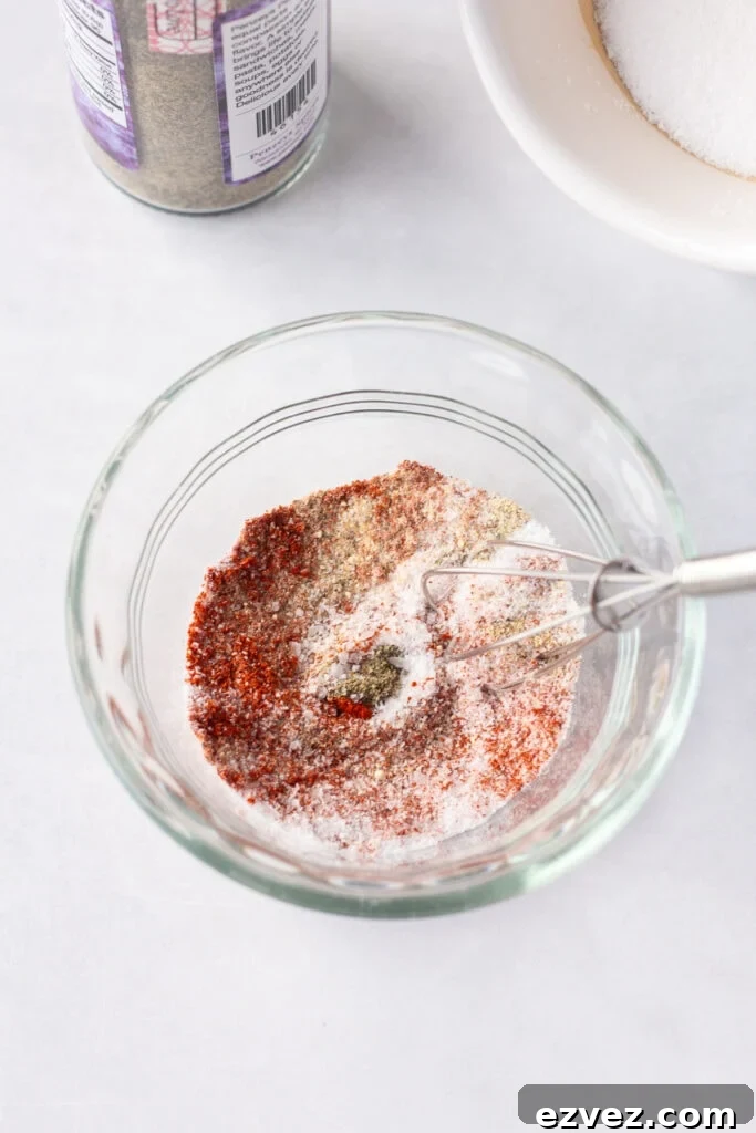 Close up of a small whisk stirring red, yellow, white, and black spices together in a small clear bowl that's on a white surface.