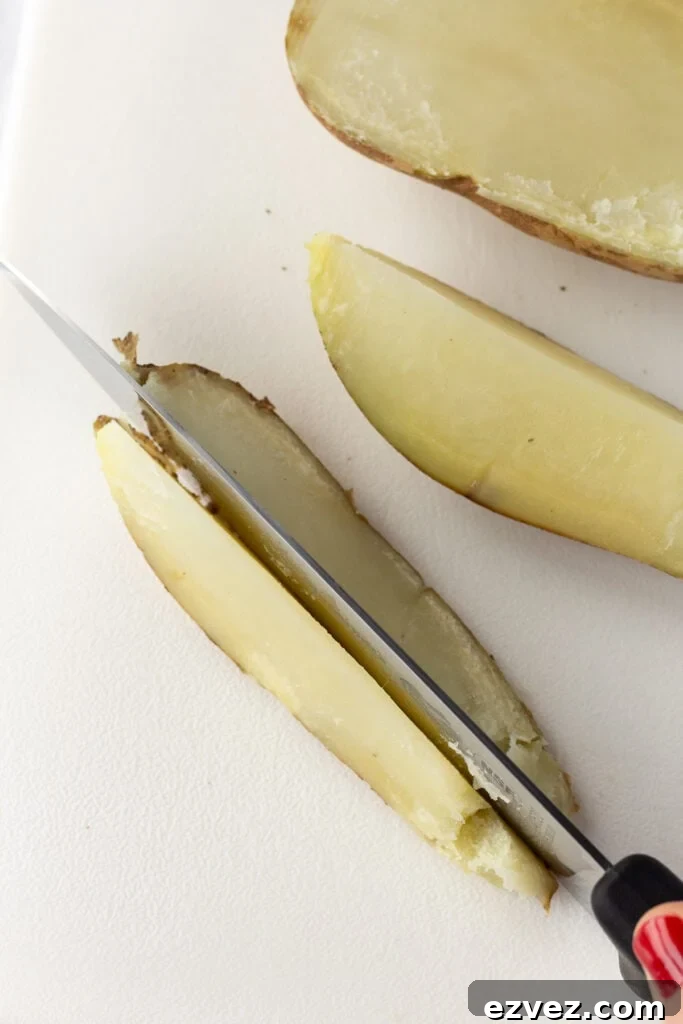 A par-cooked russet potato being cut into 8 wedge pieces on a large white cutting board with a large sharp knife.