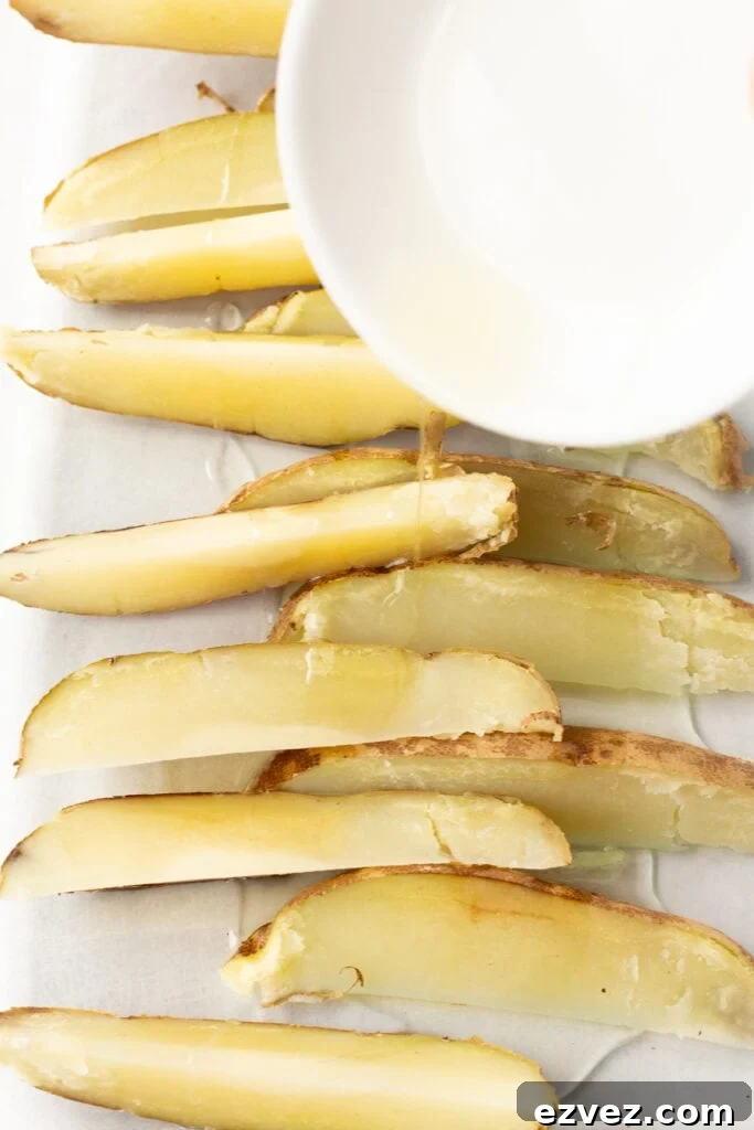 Oil being poured over par cooked russet potato wedges in a large glass dish.