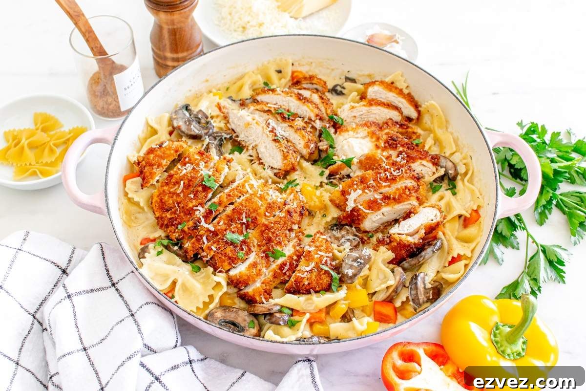 A white pan containing a dish of Louisiana Cajun chicken pasta with breaded chicken slices over bow-tie pasta, garnished with mushrooms, parsley, and grated cheese. Küchen utensils and ingredients are placed around the pan in this Cheesecake Factory copycat recipe.