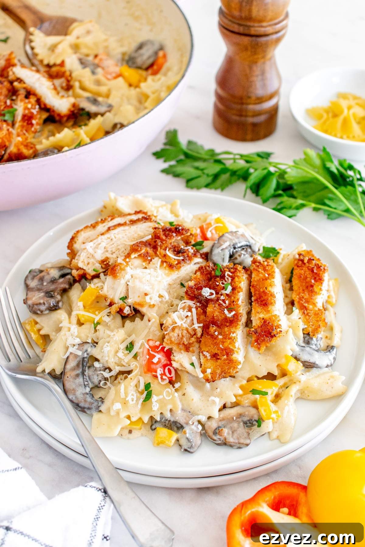 A plate of Louisiana Cajun chicken pasta with sliced breaded chicken, colorful bell peppers, mushrooms, and a light cheese garnish. This Cheesecake Factory copycat dish is served next to a fork, with a pepper grinder and fresh herbs in the background.
