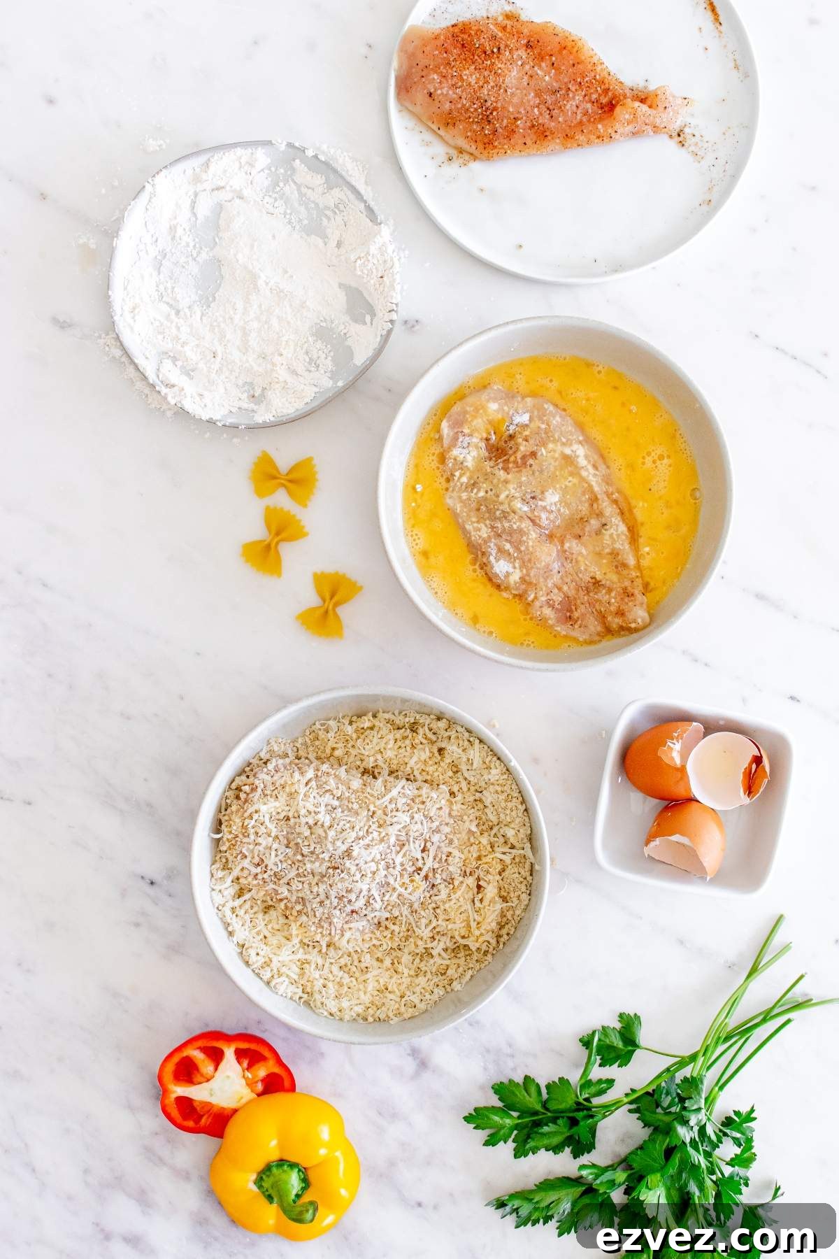 Ingredients for breaded chicken preparation, including a raw chicken breast, flour, beaten egg, breadcrumbs, half an eggshell, pasta, sliced pepper, and fresh parsley on a white surface—essential components for a Louisiana Cajun Chicken Pasta Cheesecake Factory copycat dish.