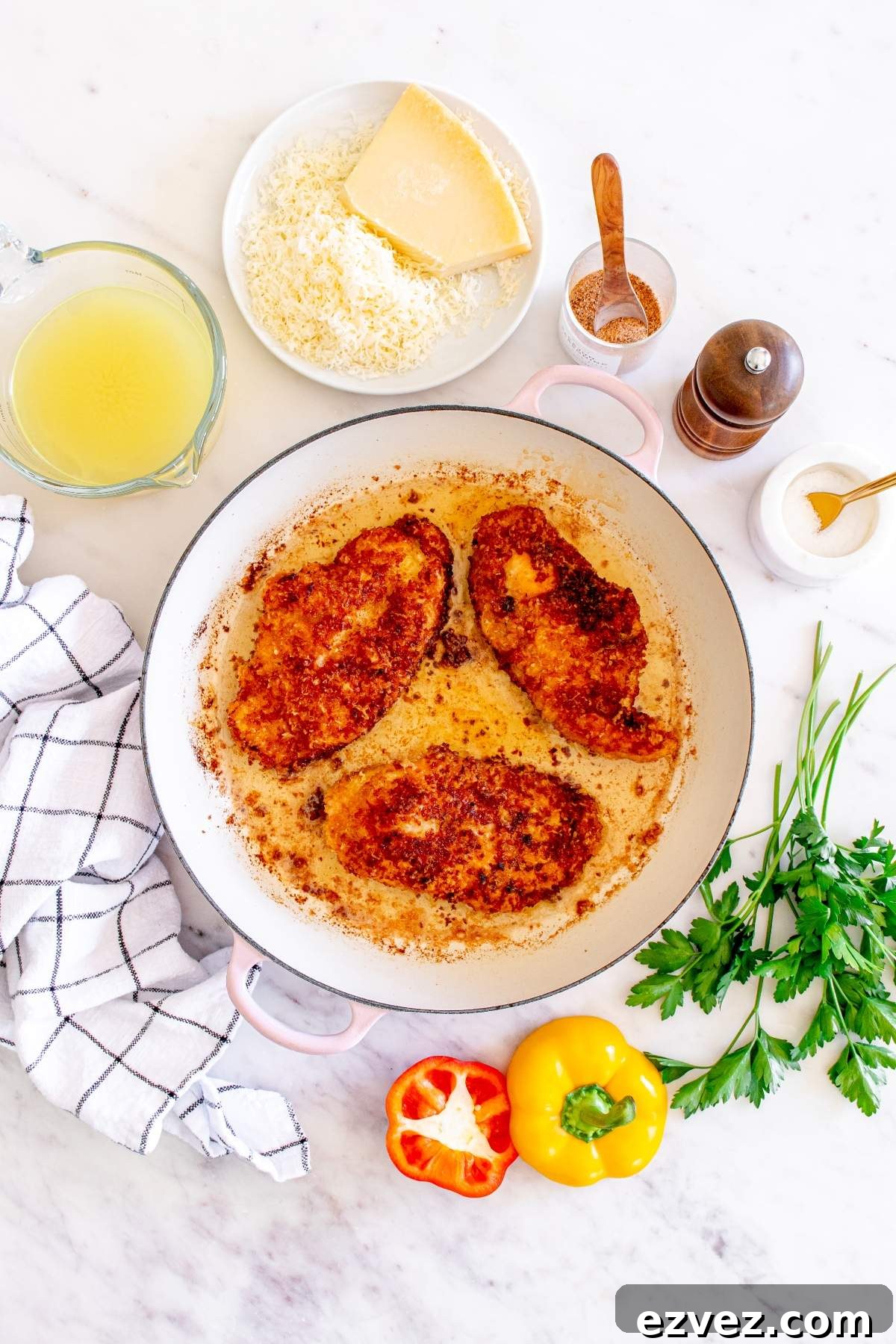 A skillet with three cooked chicken breasts sits on a counter surrounded by grated cheese, a block of cheese, seasonings, chicken broth, a striped cloth, and fresh vegetables, ready to recreate the famous Cheesecake Factory Louisiana Cajun Chicken Pasta.