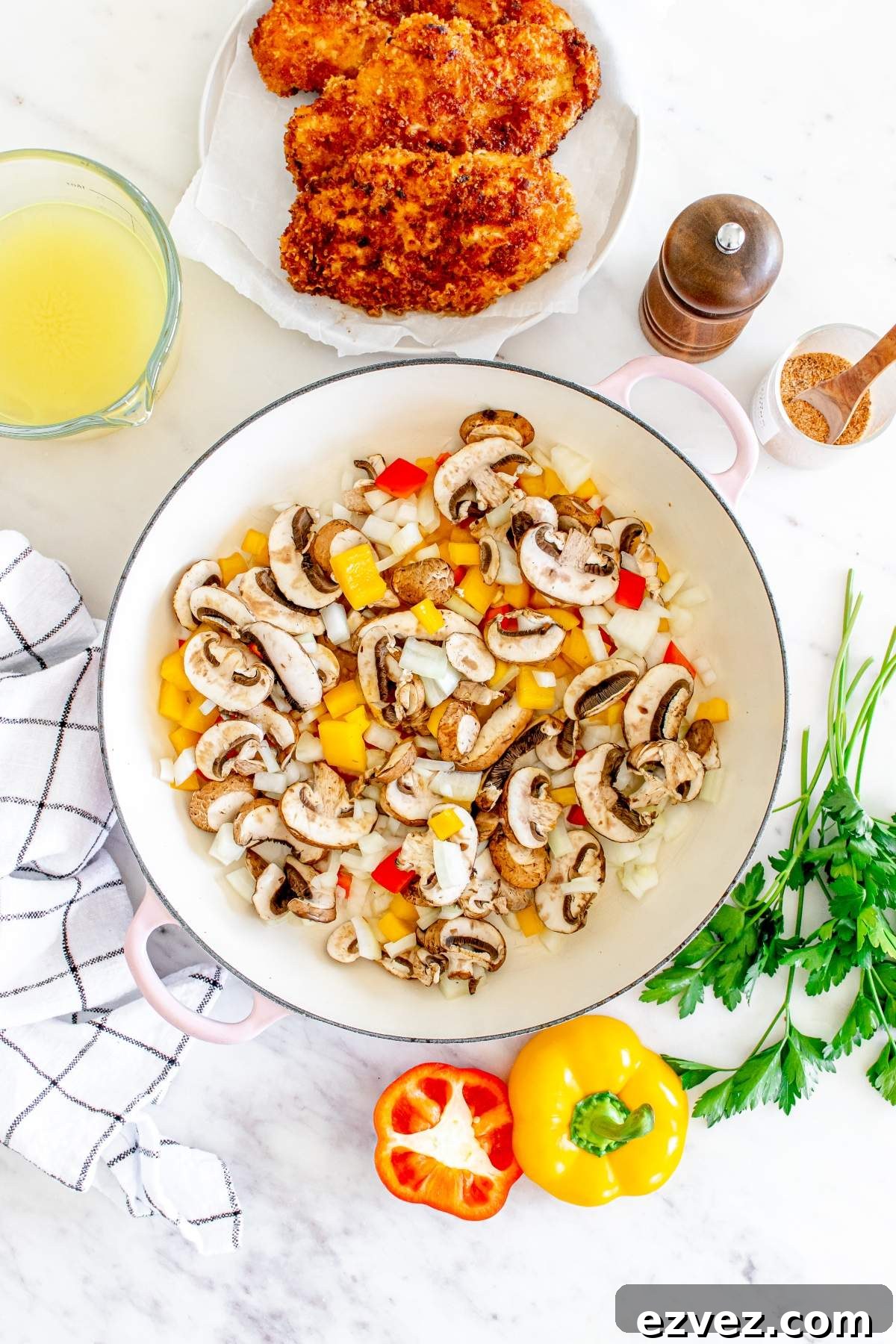 A pan with diced mushrooms, bell peppers, and onions sits on a table alongside fried chicken ready to transform into a mouthwatering Louisiana Cajun chicken pasta Cheesecake Factory copycat, complete with a pepper grinder, broth, spices, parsley, red bell pepper, and yellow bell pepper beside a cloth.