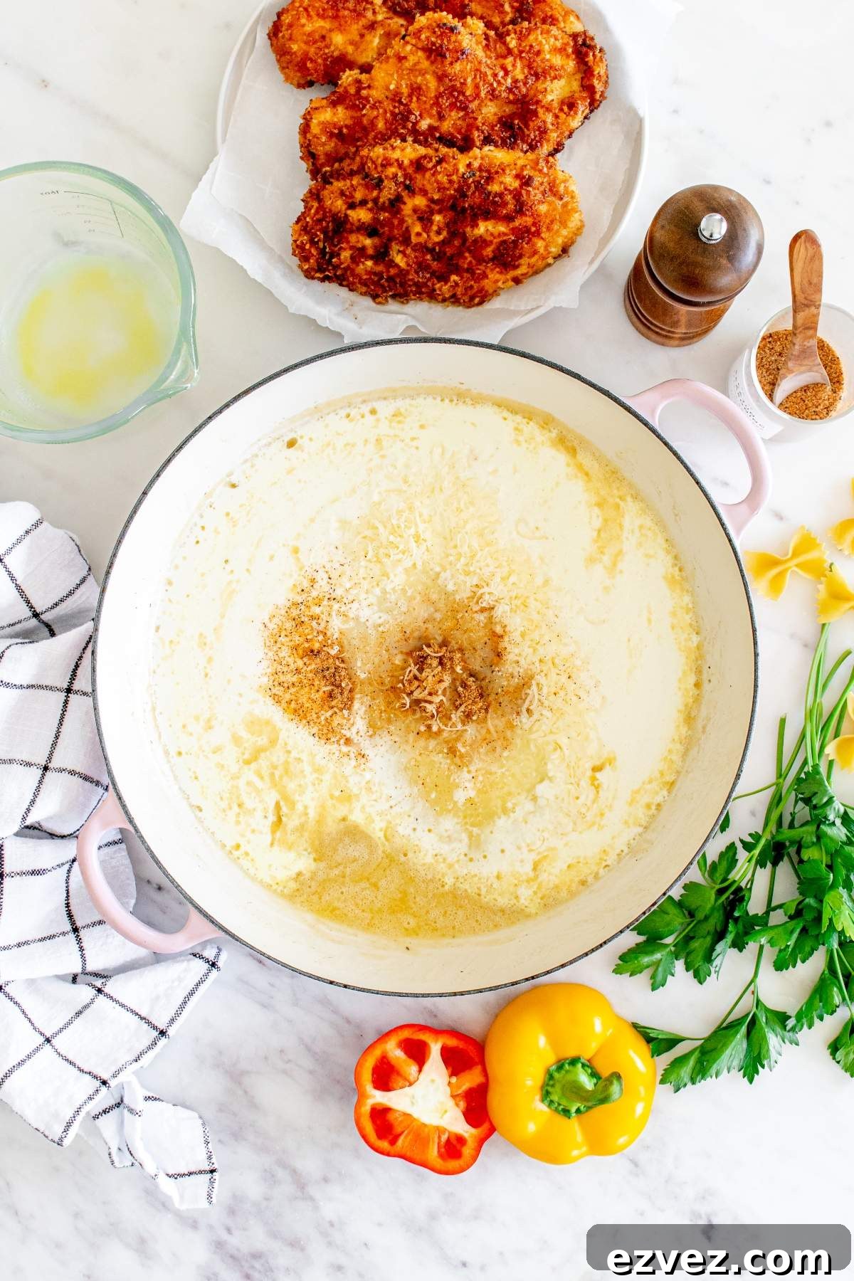 A large pot of creamy sauce with grated cheese on top, surrounded by a bell pepper, parsley, spices, a cup of melted butter, breaded chicken on a plate like the Louisiana Cajun chicken pasta from Cheesecake Factory, a pepper grinder, and a kitchen towel.