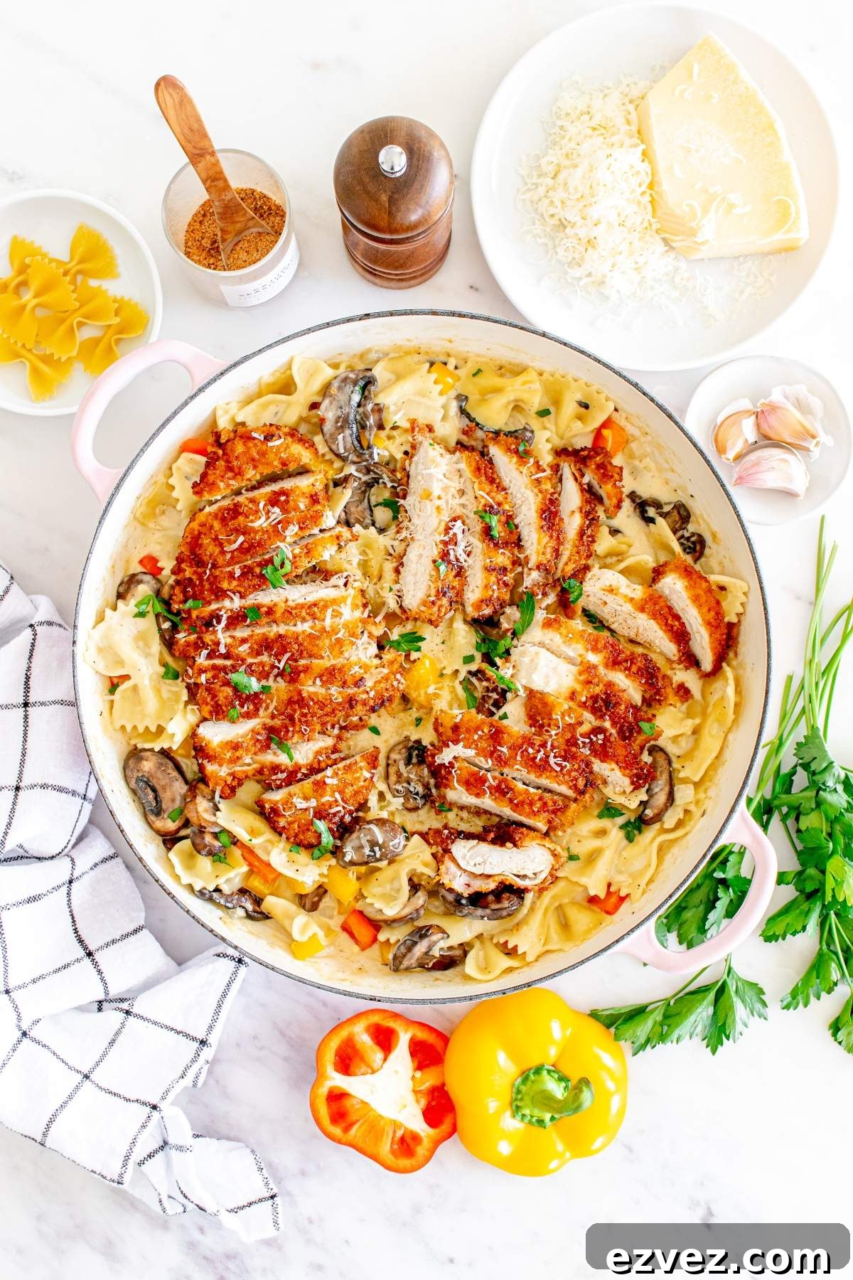 A creamy pasta dish topped with sliced, breaded, Louisiana Cajun chicken and garnished with parsley. Surrounding items include grated cheese, bell peppers, garlic, a pepper mill, and a spice bowl on a white surface for that perfect Cheesecake Factory copycat experience.