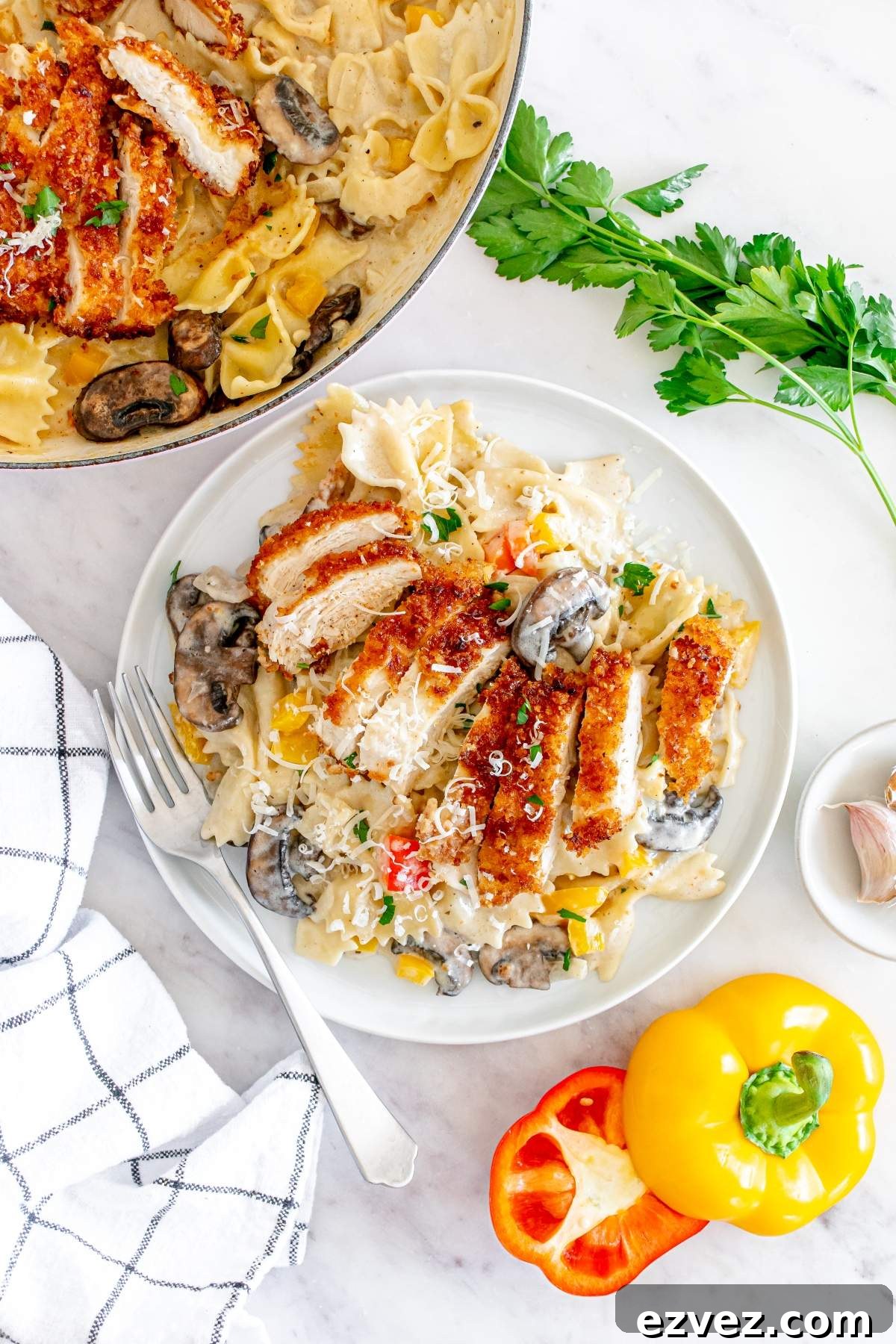 A plate of Louisiana Cajun chicken pasta topped with crispy chicken slices, mushrooms, and vegetables beside fresh parsley, bell peppers, and a fork. A striped cloth napkin is placed nearby on a white surface in this Cheesecake Factory copycat dish.