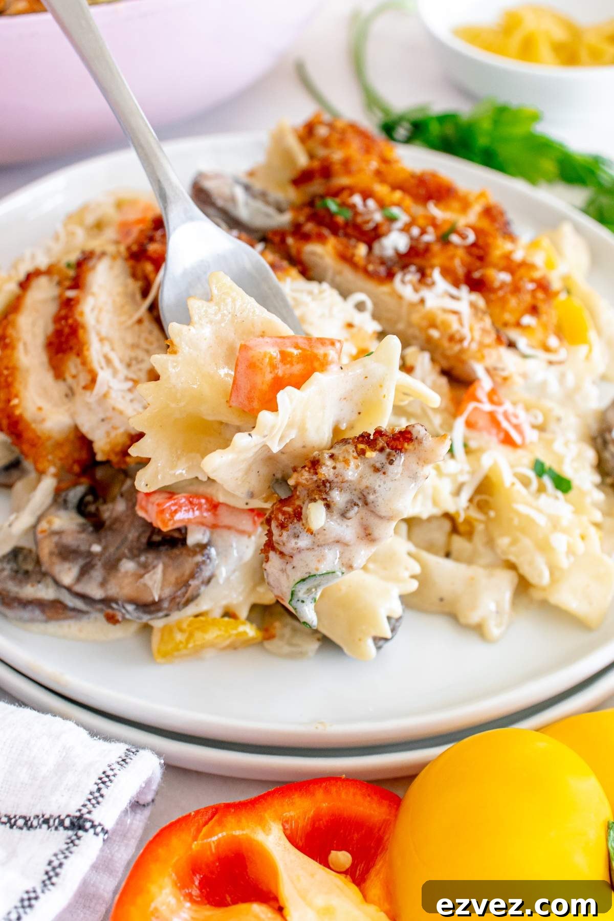 A fork picking up a piece of bowtie pasta from a plate containing pasta with vegetables and creamy sauce, garnished with fresh herbs, accompanied by crispy Louisiana Cajun chicken pieces on the side. Peppers visible in the foreground.