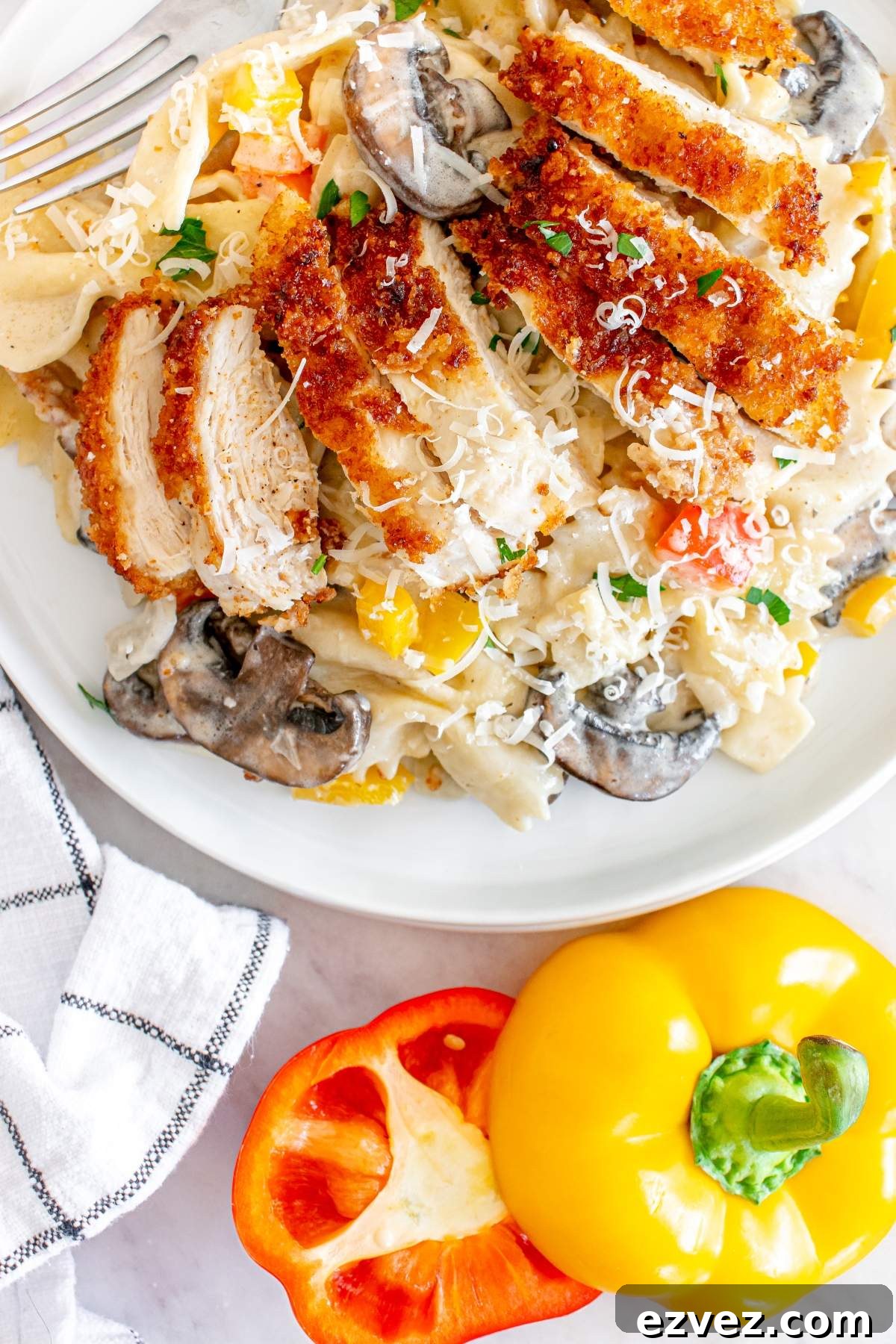 A plate of Louisiana Cajun chicken pasta, topped with sliced breaded chicken, mushrooms, and shredded cheese. A fork rests on the plate, and a cut yellow bell pepper and a half-cut red bell pepper are beside it in this Cheesecake Factory copycat dish.