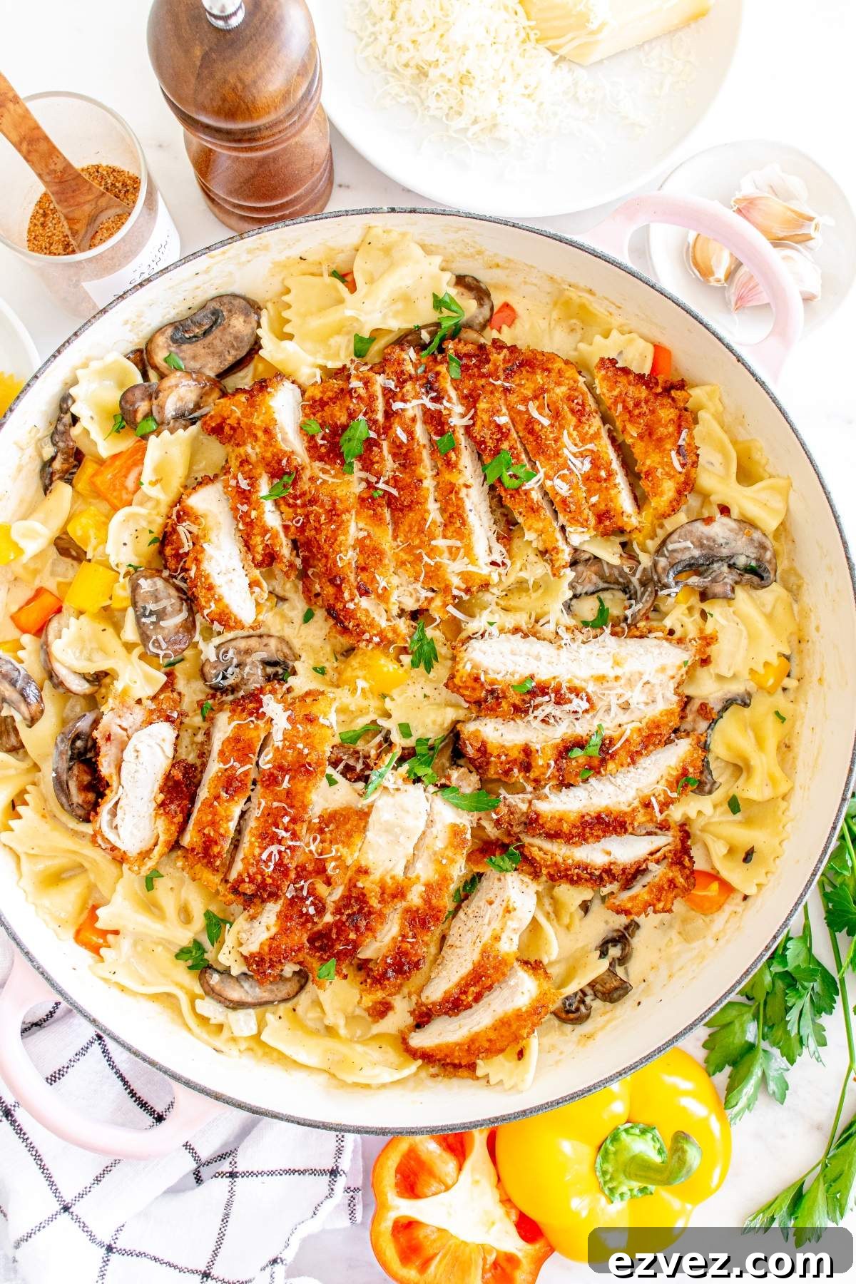 A skillet filled with bow-tie pasta in a creamy sauce, topped with slices of breaded Louisiana Cajun chicken, mushrooms, and garnished with parsley. Adjacent are a pepper grinder, cheese, garlic, bell peppers, and a cloth—just like the Cheesecake Factory copycat version.