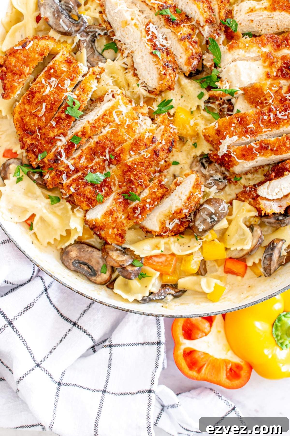 A skillet containing Louisiana Cajun chicken pasta with breaded chicken slices, mushrooms, and bell peppers, garnished with grated cheese and parsley, alongside a checkered napkin and half of a yellow bell pepper. This dish is a Cheesecake Factory copycat sure to impress.