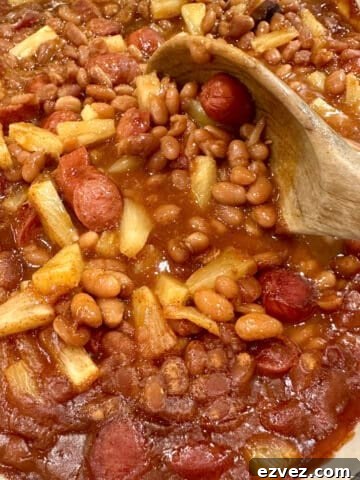 Baked beans, franks and pineapple pieces in a baking dish with a spoon.