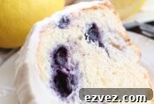A slice of lemon blueberry bundt cake frosted with lemon cream cheese frosting sitting on a small white plate. Above the plate is a whole lemon and a blueberry lemon bundt cake frosted with lemon cream cheese frosting with a slice taken out of it.