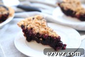 a slice of blueberry pie with a crumble topping on a white plate. A second slice is in the background and you can also see a corner of the pie pan the slices came out of.