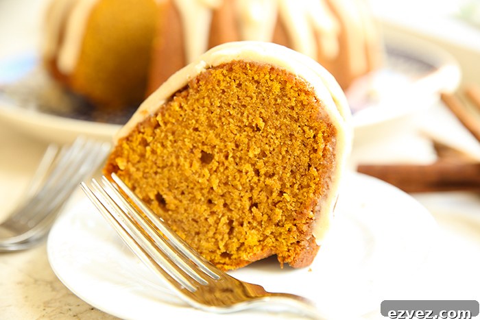 A piece of pumpkin spice bundt cake with frosting and a fork on a dessert plate. Faded in the background is a pumpkin bundt cake with brown butter frosting with one slice cut out of it.