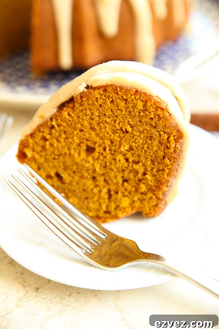 A slice of pumpkin spice cake on a small white plate with a fork. Faded in the background is a pumpkin bundt cake with a slice cut out of it.