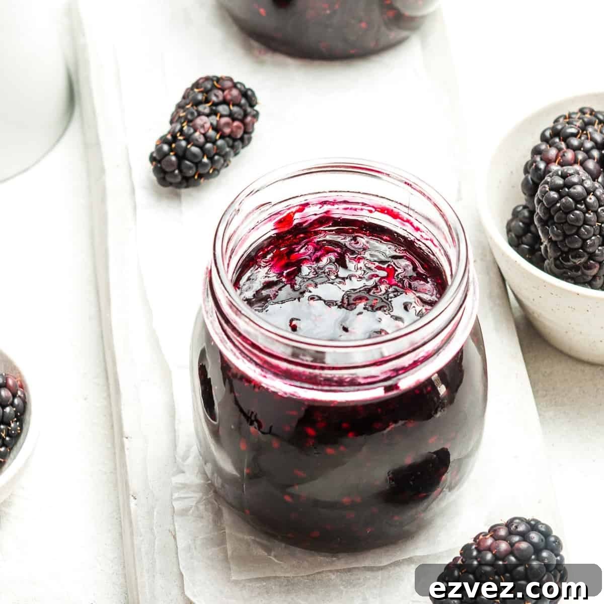Open jar of jam, sitting on some white paper, with some blackberries around the edge.
