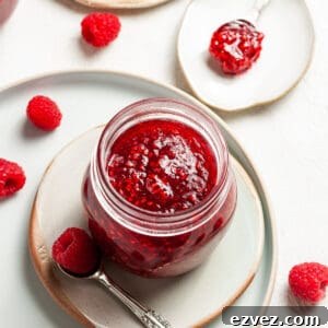 Overhead shot of open jar of jam, sitting on a plate, with a spoon and some raspberries on the edge.