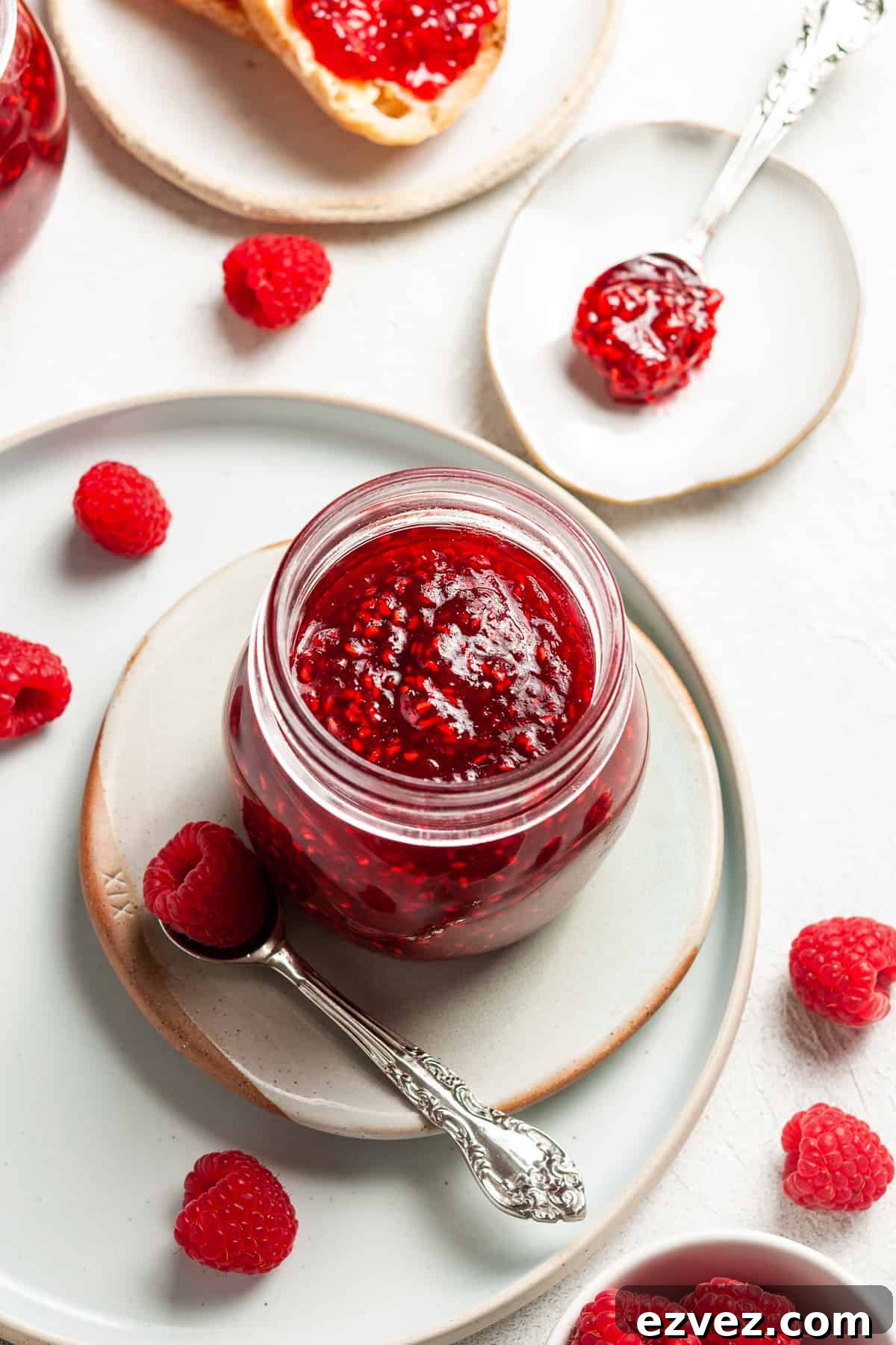 Overhead shot of open jar of jam, sitting on a plate, with a spoon and some raspberries on the edge.