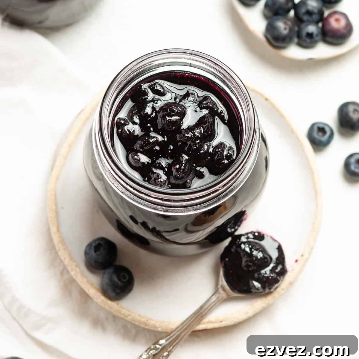 Overhead shot of open jar of jam, sitting on a plate, with a spoon and some blueberries on the edge.