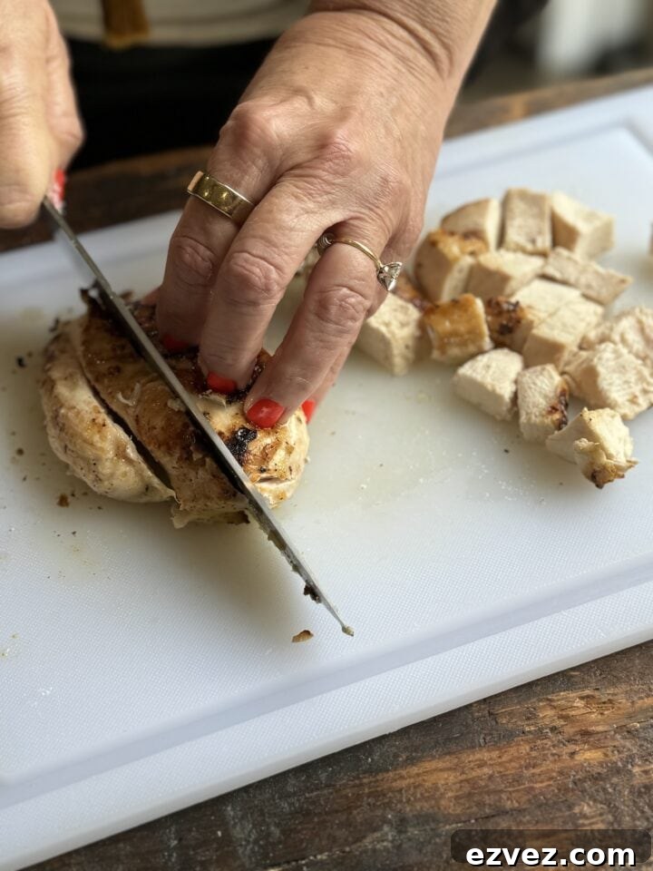 two hands with red nail polish and gold rings cutting chicken breast into cubes