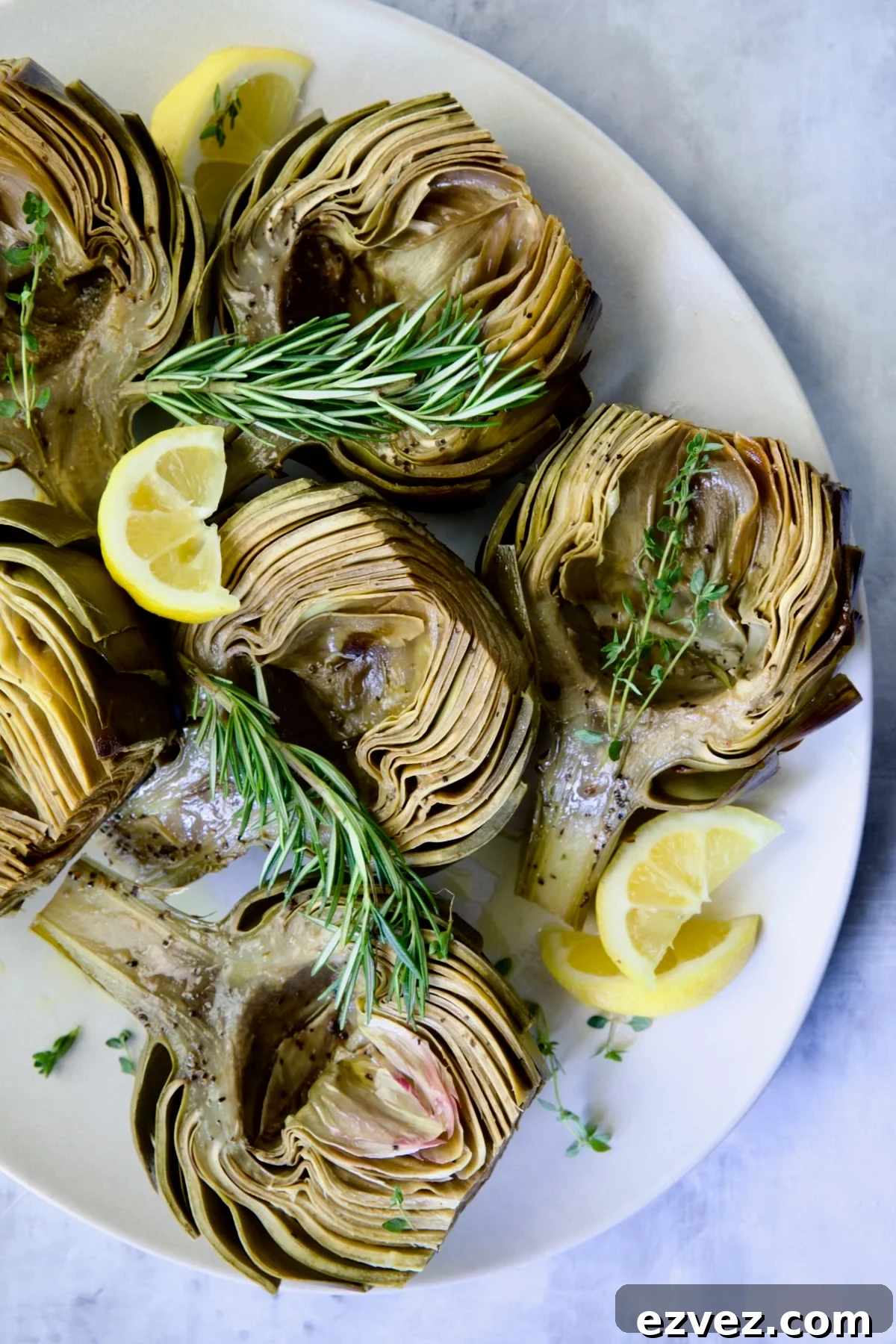 Roasted Artichokes with Garlic Butter on white oval platter garnished with lemon wedges and herb sprigs.