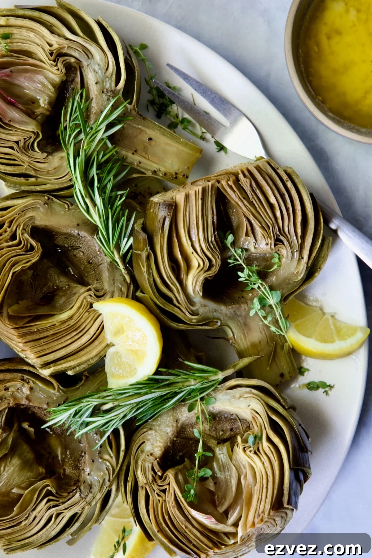 Roasted Artichokes with Garlic Butter on white oval platter garnished with herb sprigs and lemon wedges.