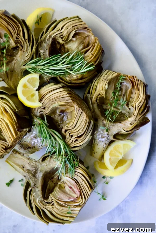Roasted Artichokes with Garlic Butter on white oval platter garnished with lemon wedges and herb sprigs.