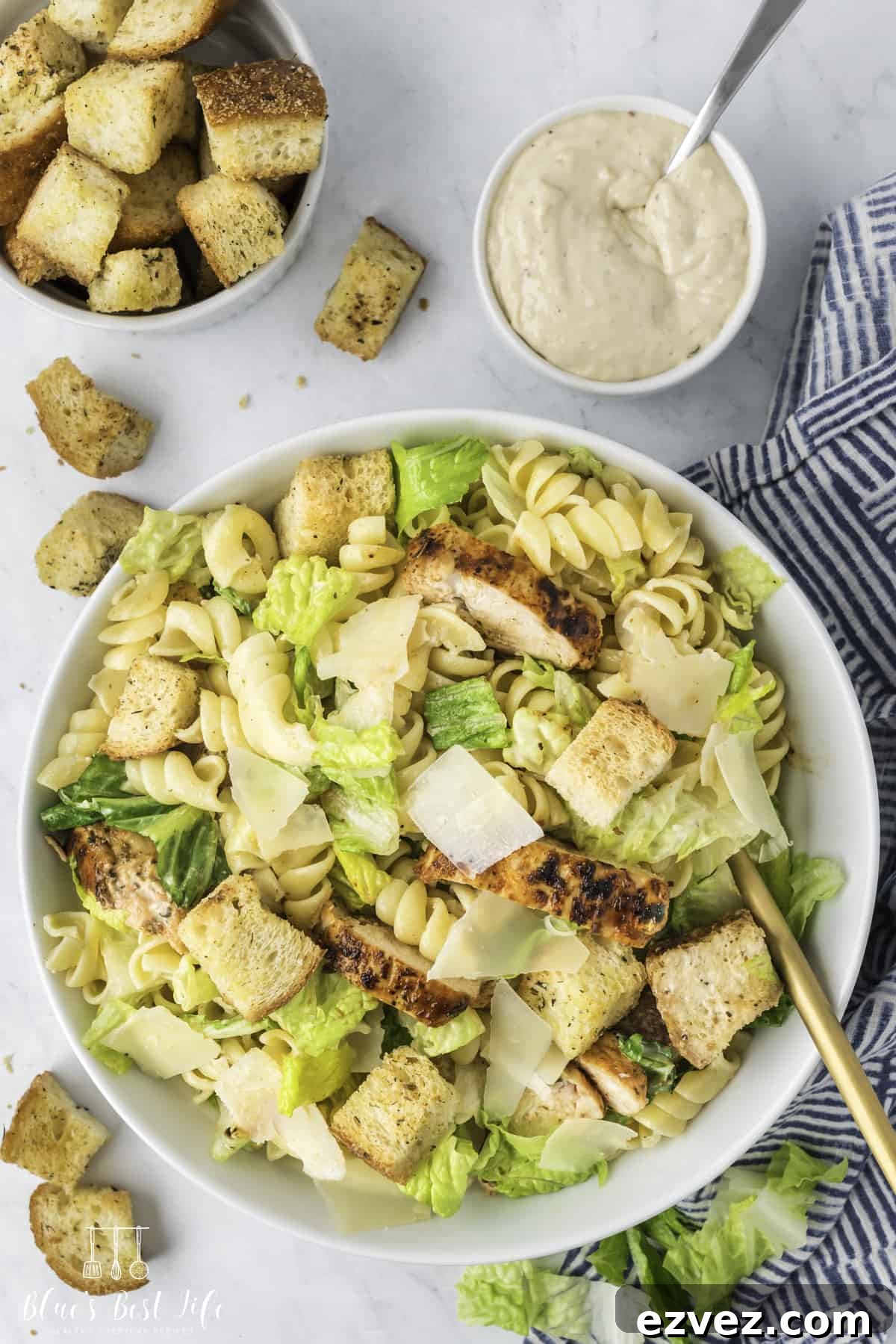 Top shot of a bowl with the chicken Caesar pasta salad, a small bowl of Caesar dressing and a bowl of croutons on the side. 