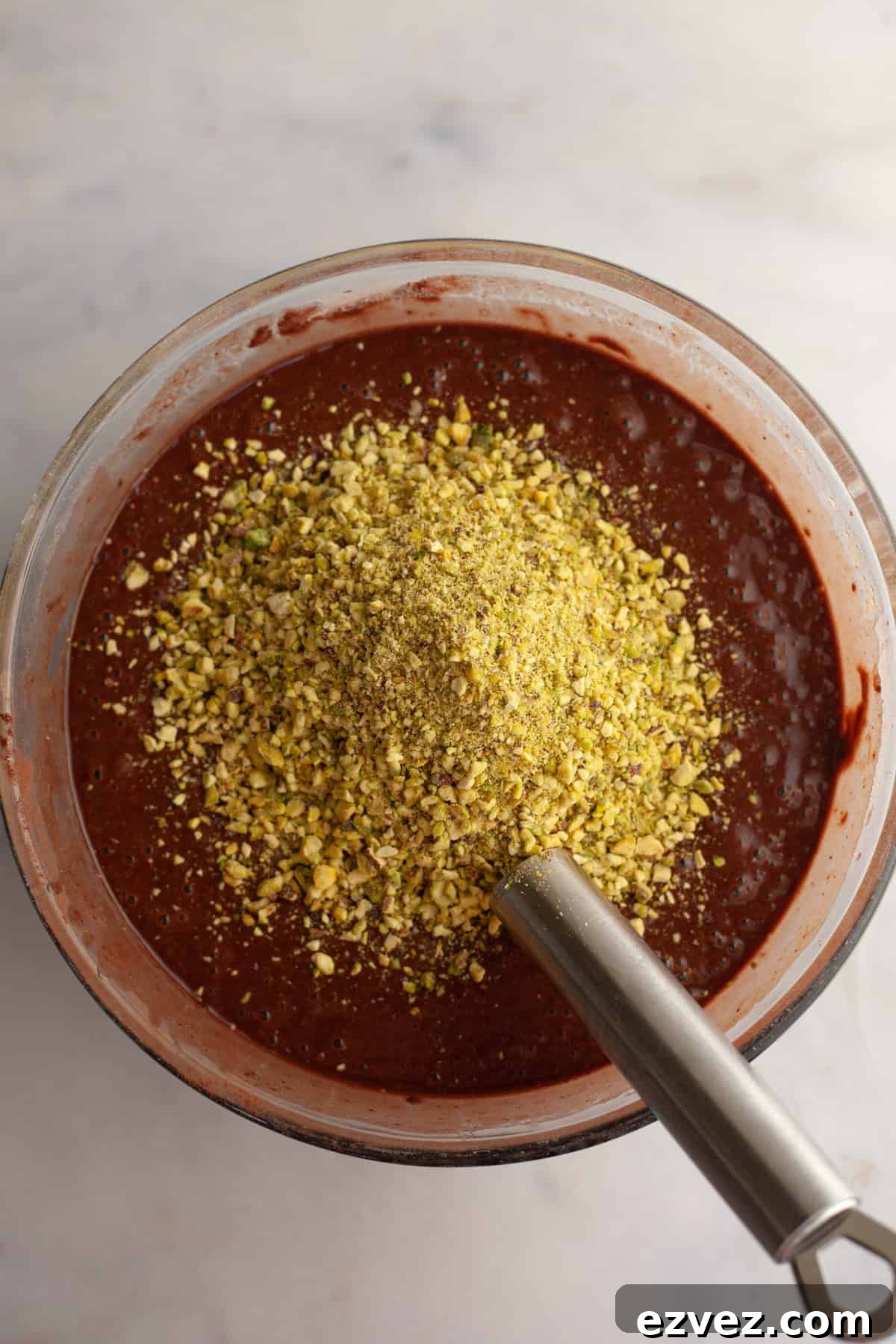 Chocolate cake batter with crushed pistachios, with a silver whisk in a glass bowl.