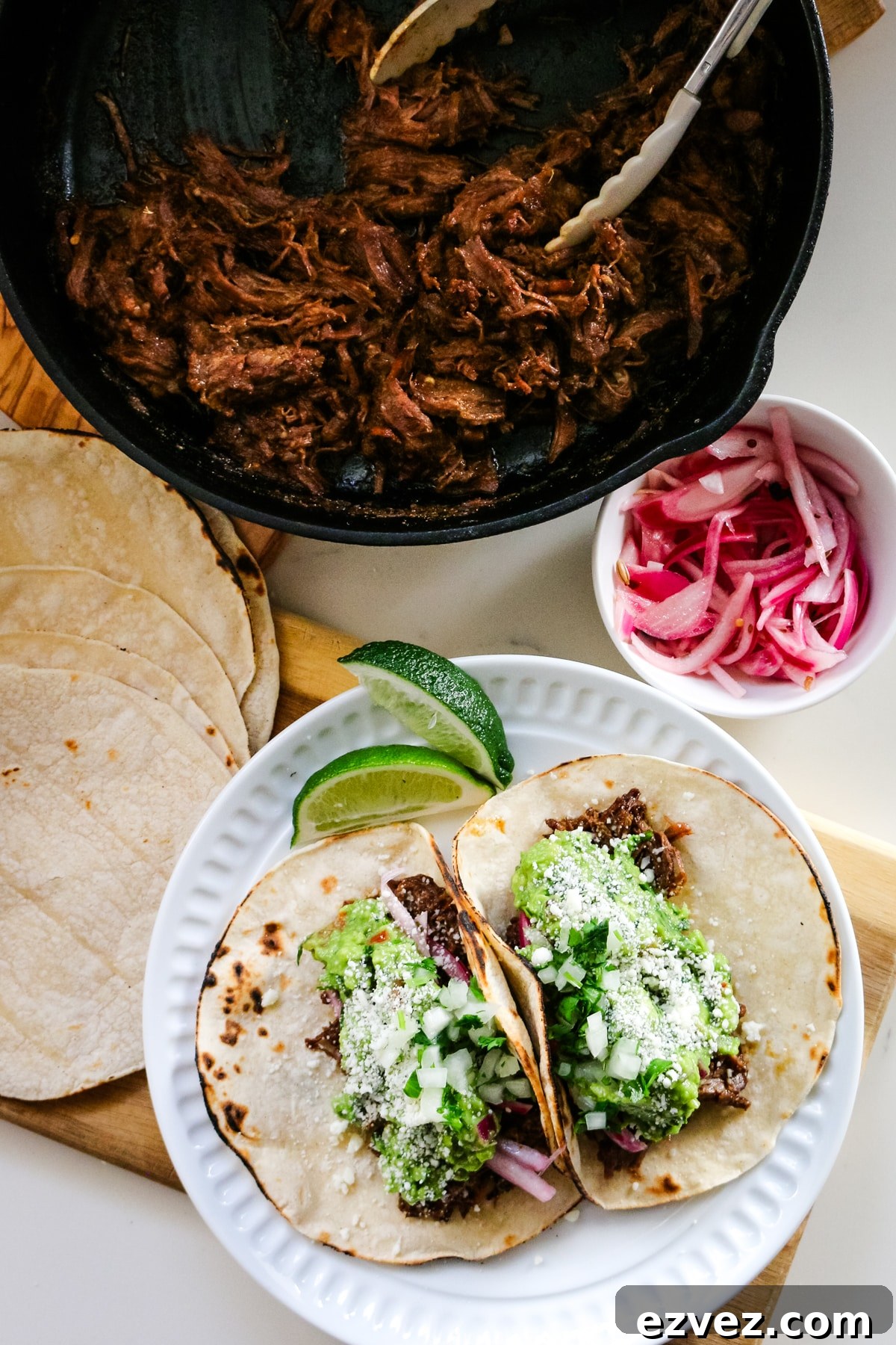 Slow-Cooker Shredded Beef Tacos for Flavorful Weeknight Meals 2 Roast tacos on a white plate with roasted beef in a pan, with tacos and onions beside it.