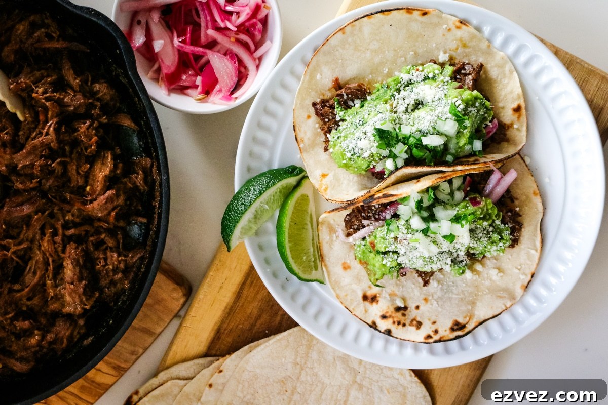 Slow-Cooker Shredded Beef Tacos for Flavorful Weeknight Meals 9 Roast tacos on a white plate with roasted beef in a pan, with tacos and onions beside it.