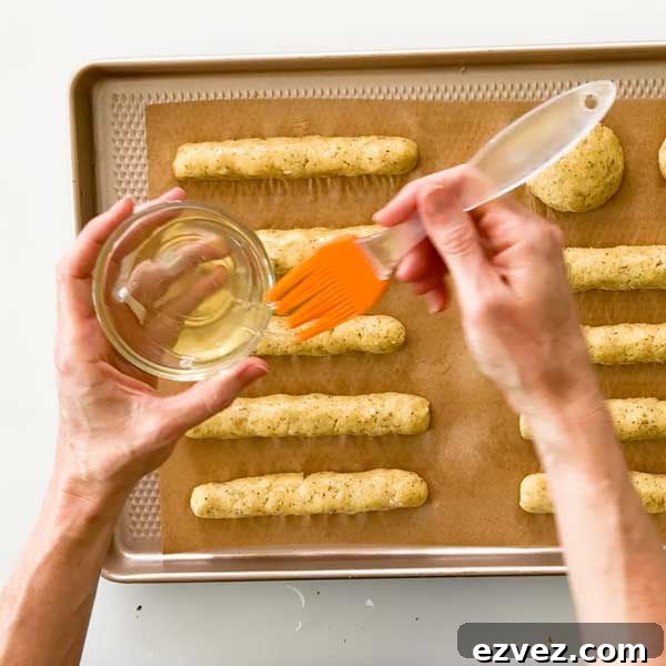 Brushing egg white on the shaped bread sticks with a pastry brush on a  baking sheet.