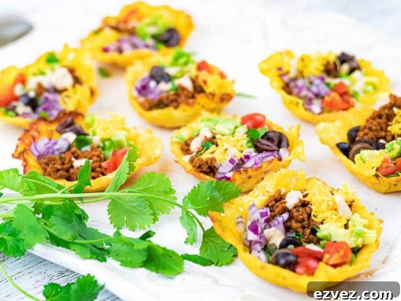 11 Party Appetizers Guests Devour Immediately 4 Mini taco cups filled with ground meat, cheese, lettuce, tomatoes, olives, and onions are arranged on a white surface with fresh cilantro in the foreground.