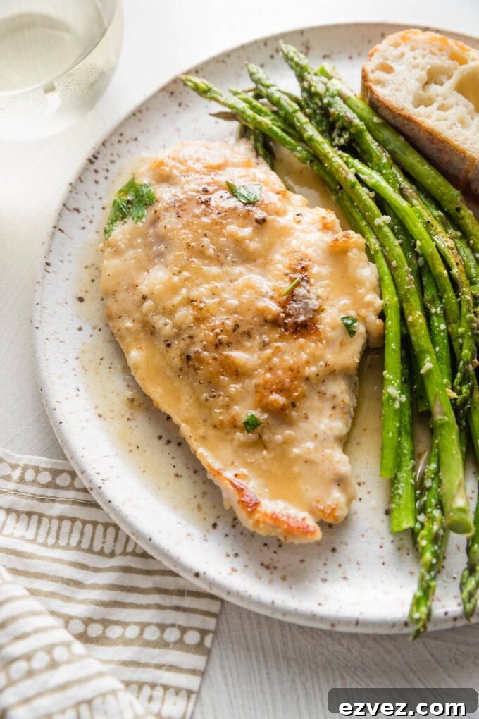 Small white plate with a tender pan-fried chicken breast in white wine sauce, roasted asparagus, and a slice of bread.