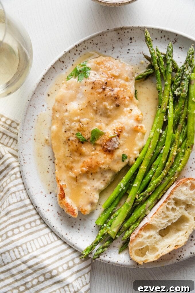 Small white plate with a tender pan-fried chicken breast in white wine sauce, roasted asparagus, and a slice of bread.