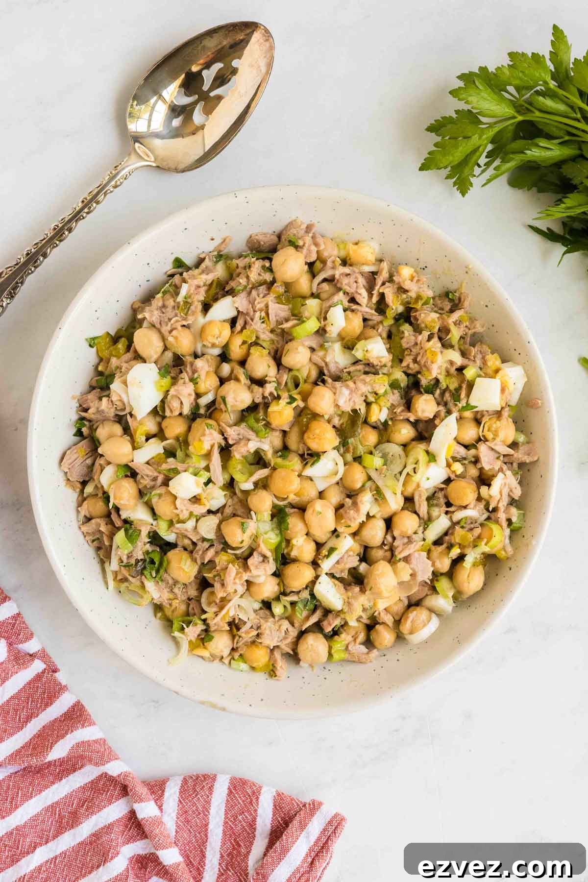A bowl of chickpea and tuna salad with chopped vegetables, next to a serving spoon, a red-striped towel, and a sprig of parsley on a white surface.