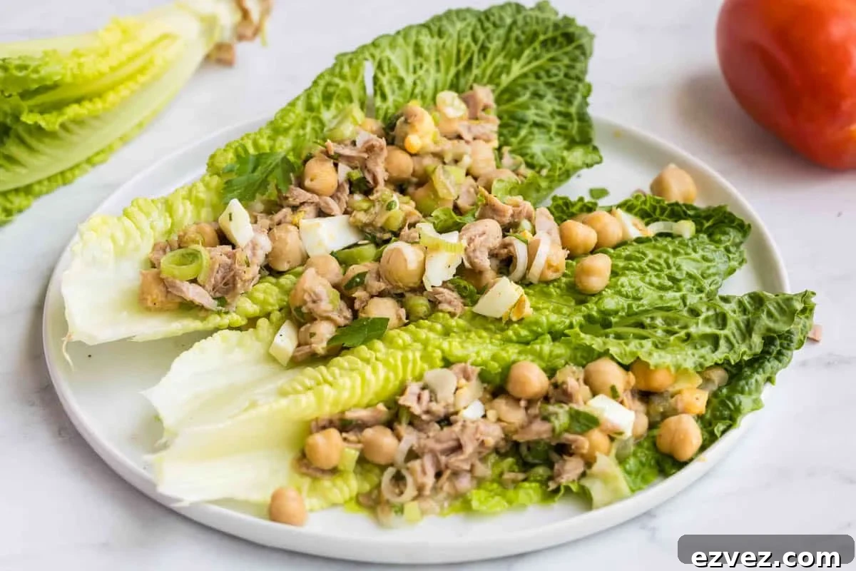 Three romaine lettuce leaves on a white plate, topped with a mixture of chickpeas, tuna, chopped egg, and green onions. A tomato and more lettuce are in the background.