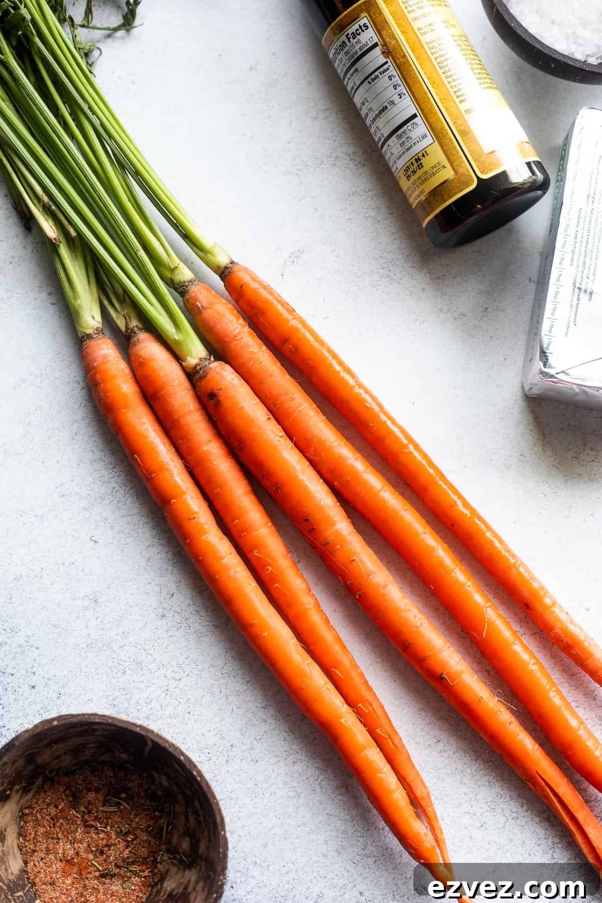 Give long carrots with green stalks arranged on a white background with balsamic vinegar and a seasonings mixture placed around the carrots.