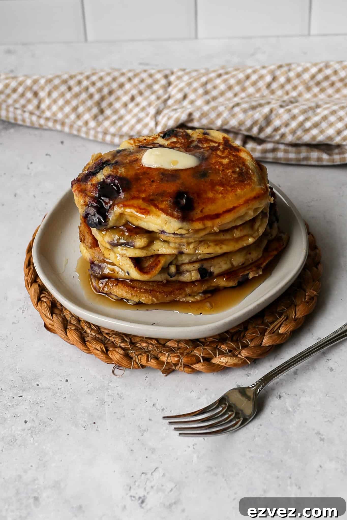 a white plate of five gluten free high protein lemon blueberry pancakes on a woven trivet next to a tan gingham kitchen towel