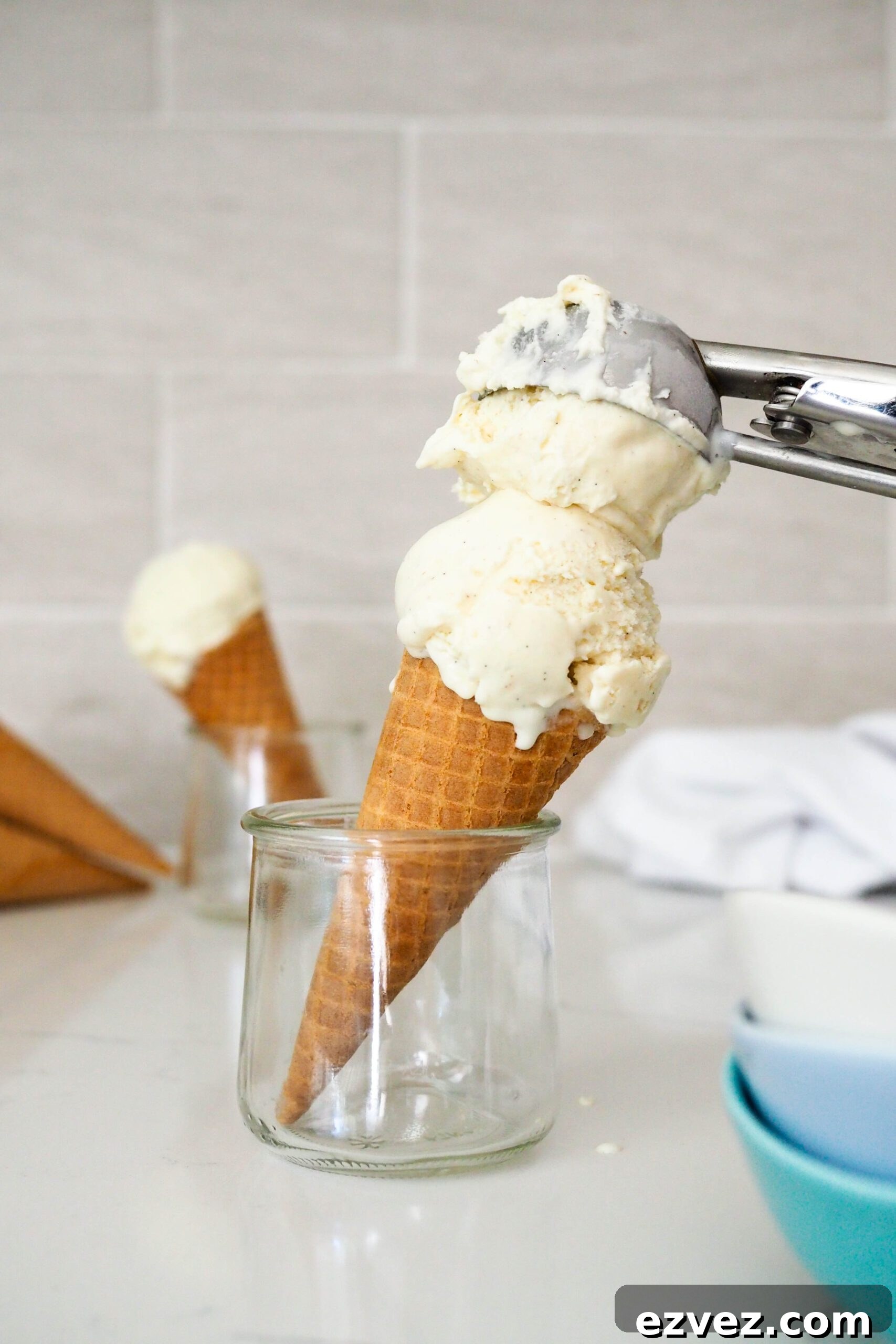 An ice cream scoop adds a second scoop to a sugar cone.